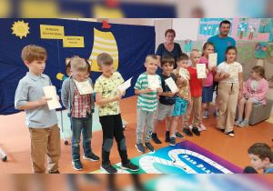 Ośmioro dzieci wyróżnionych w konkursie z dyplomami i nagrodami książkowymi stoi na tle wakacyjnej dekoracji.