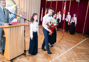 Delegacja uczniów niesie kwiaty pod tablicę pamiątkową Patrona.